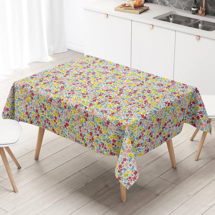 Bunte Wachstuch-Tischdecke mit dichtem Streublumenmuster in Rot, Gelb und Rosa sowie grünen Blättern auf hellem Hintergrund, über eine rechteckige Tischkante drapiert.