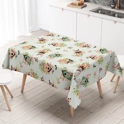Wachstuch-Tischdecke mit Vogelhäuschen, kleinen Vögeln, Schmetterlingen und rosa Blüten auf hellem mintfarbenem Hintergrund. Grüne Blätter und natürliche Holztöne verleihen dem verspielten Frühlingsdesign eine frische, freundliche Ausstrahlung.