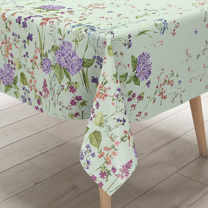 Wachstuchtischdecke mit romantischem Blumenmuster auf mintgrünem Hintergrund. Zarte Wildblumen, filigrane Zweige und Blütenranken in Rosa, Blau, Lila und Rot verteilen sich locker über die Fläche. Große violette Hortensien setzen dekorative Akzente entlang der oberen und unteren Bereiche. Das detailreiche, aquarellartige Dessin wirkt leicht, natürlich und verspielt und verleiht dem Tisch eine frische, florale Ausstrahlung.
